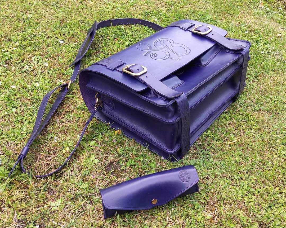 Matching messenger bag and pouch hand-made from a vibrant purple blue leather with gold hardware, placed outside on a lawn.