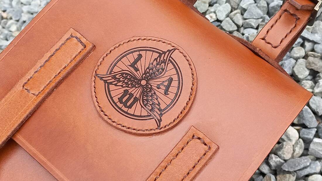 A bag hand-made from light brown leather with visible stitched, stamped with a black circular emblem featuring a wheel, three wings and the letters L, A and W.