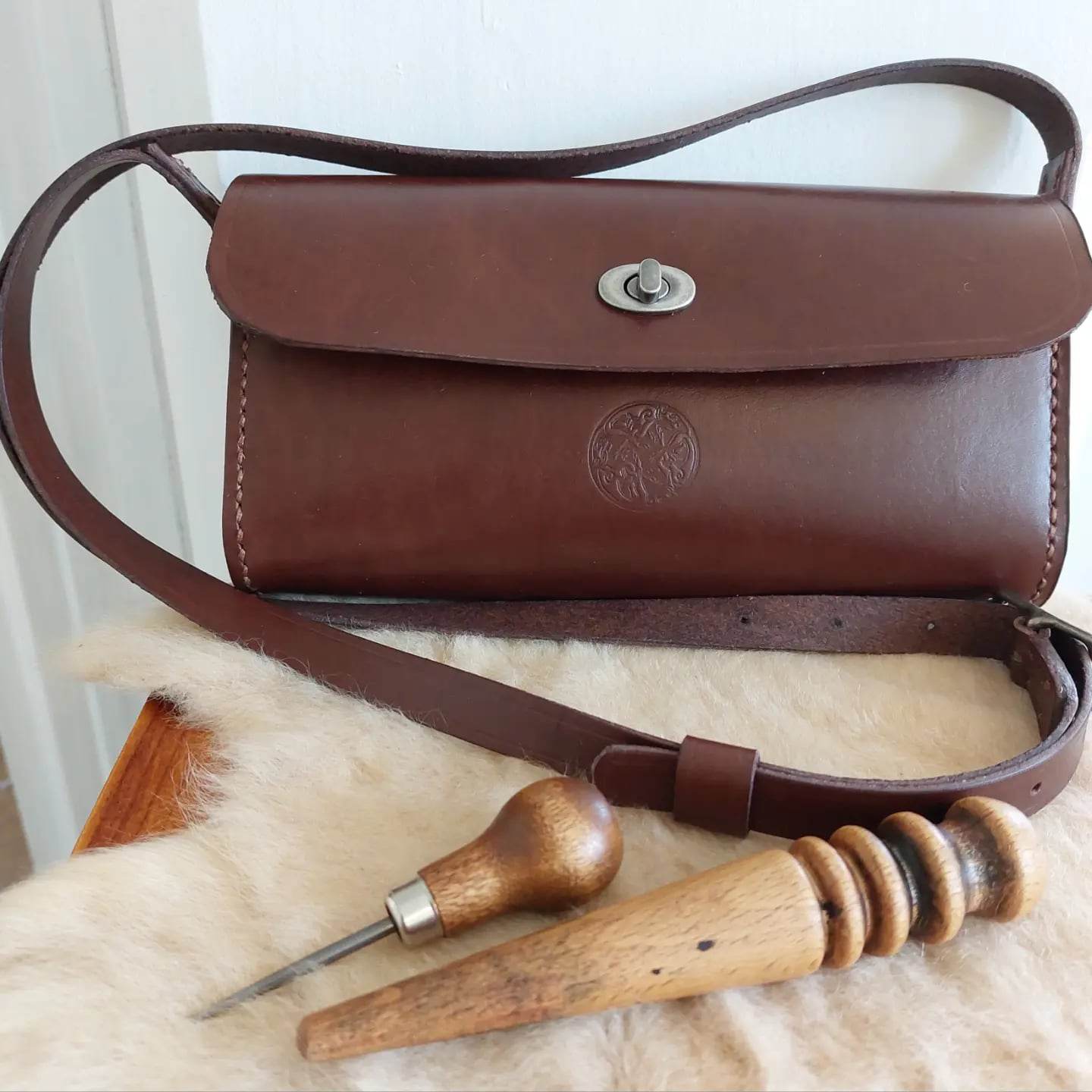 Handbag with shoulder straps hand-made with light brown leather with silver hardware and stamped with the Albannach Crafts logo. Placed atop a sheepskin alongside some leather-working tools.
