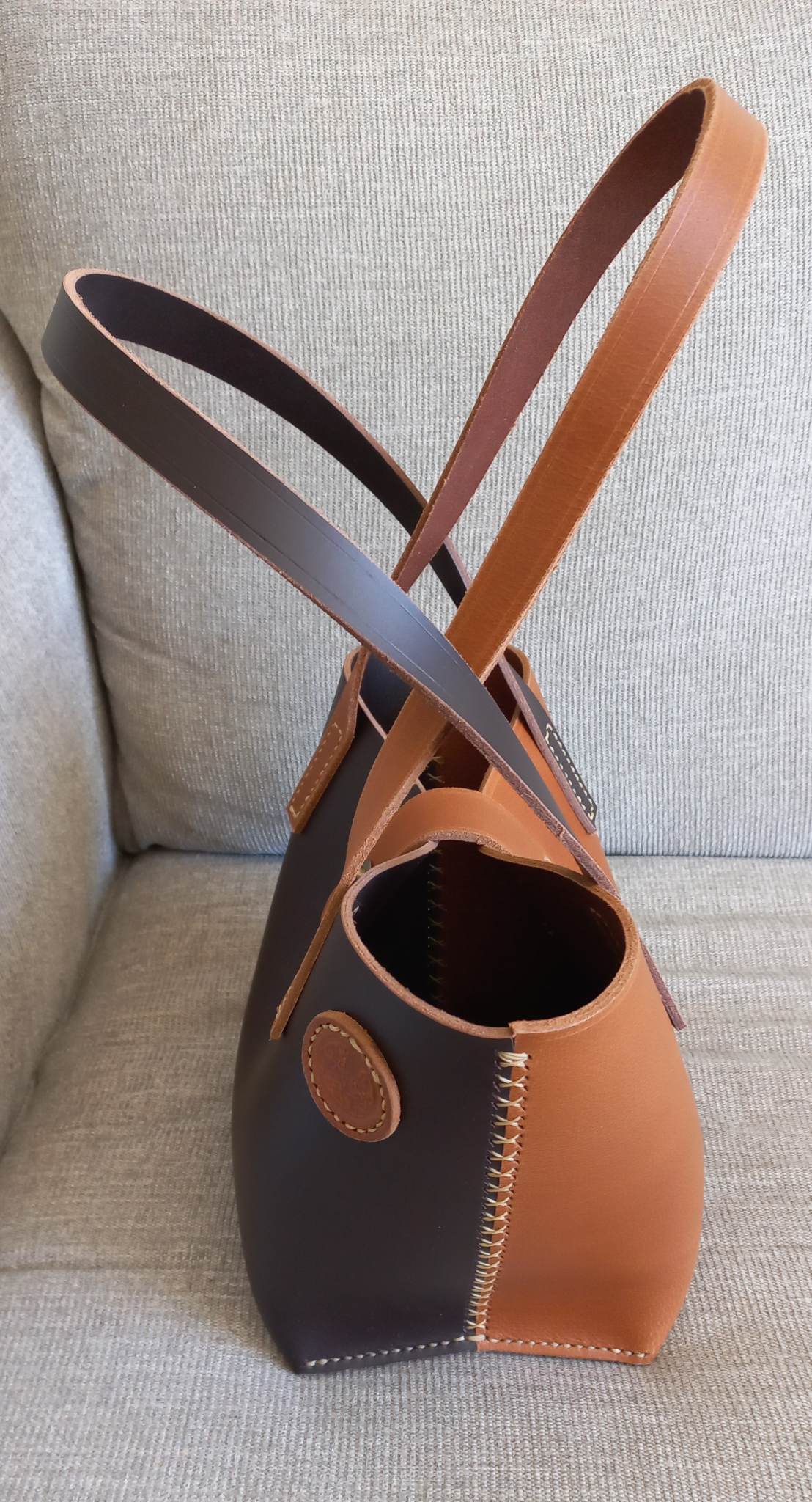 A two-tone tote bag hand-made with black and light brown leather, with circular detailing and visible stitching, placed atop an armchair.