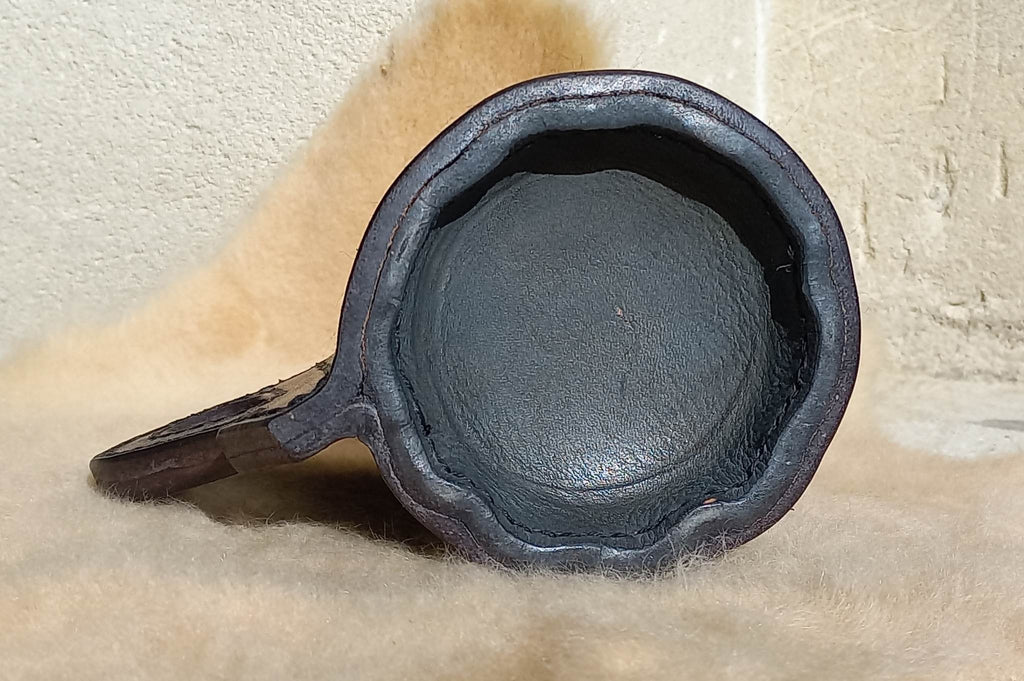 The bottom of a Replica 17th Century Blackjack made from quality bullock hide from Dunedin, complete with handle, sitting atop a wool carpet.