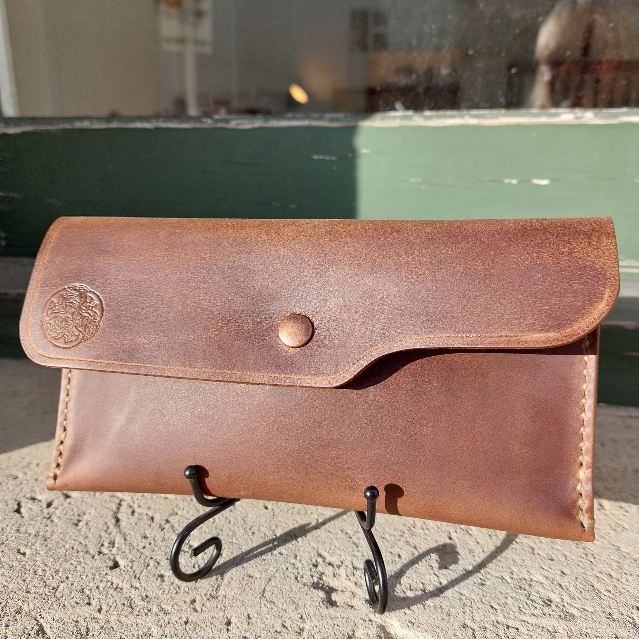 A wallet pouch hand-made with light brown leather, stamped with the Albannach Crafts logo. Held upright by a metal stand.