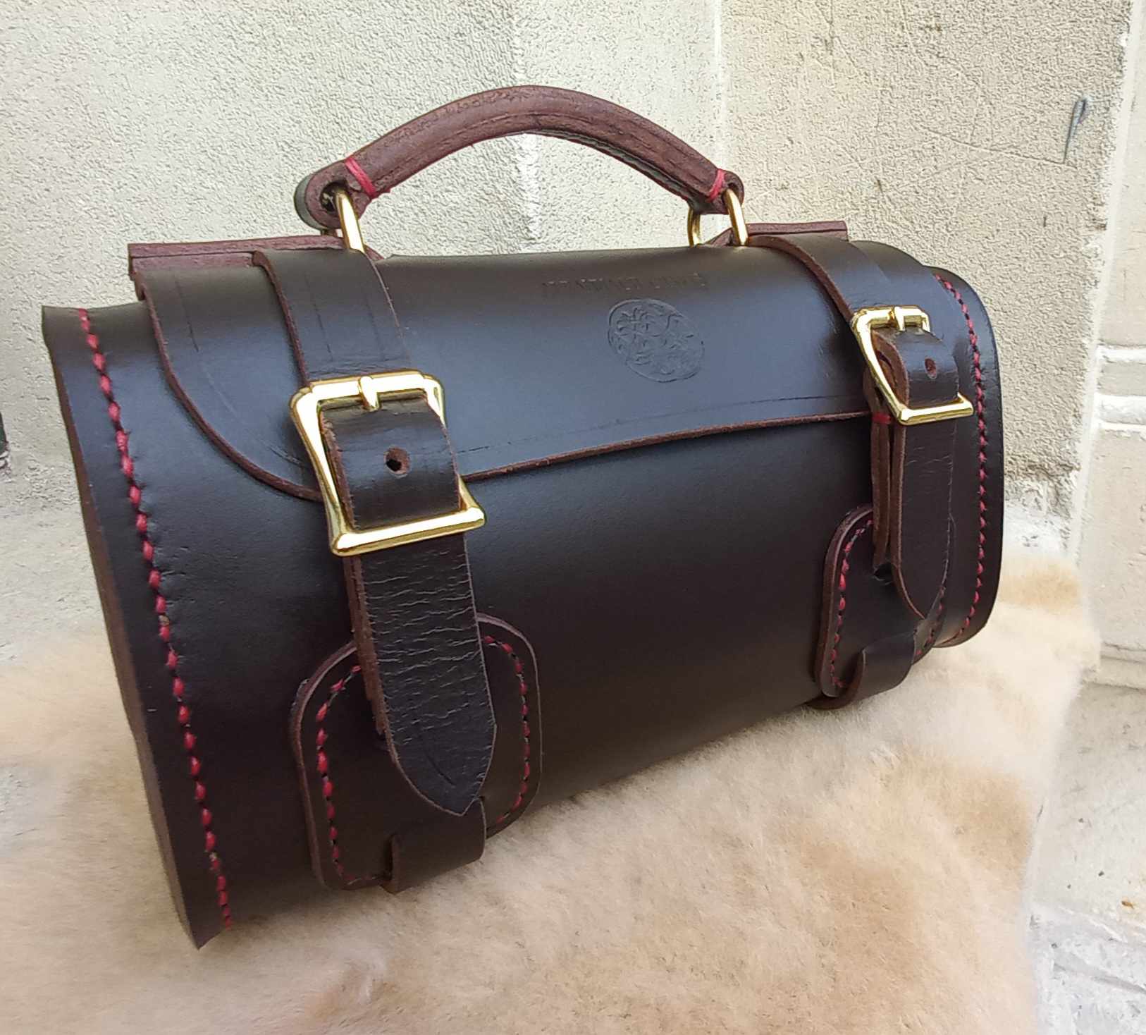 A sportsbag designed to hold petanque balls, hand-made with dark brown leather and gold hardware, placed atop a sheepskin.