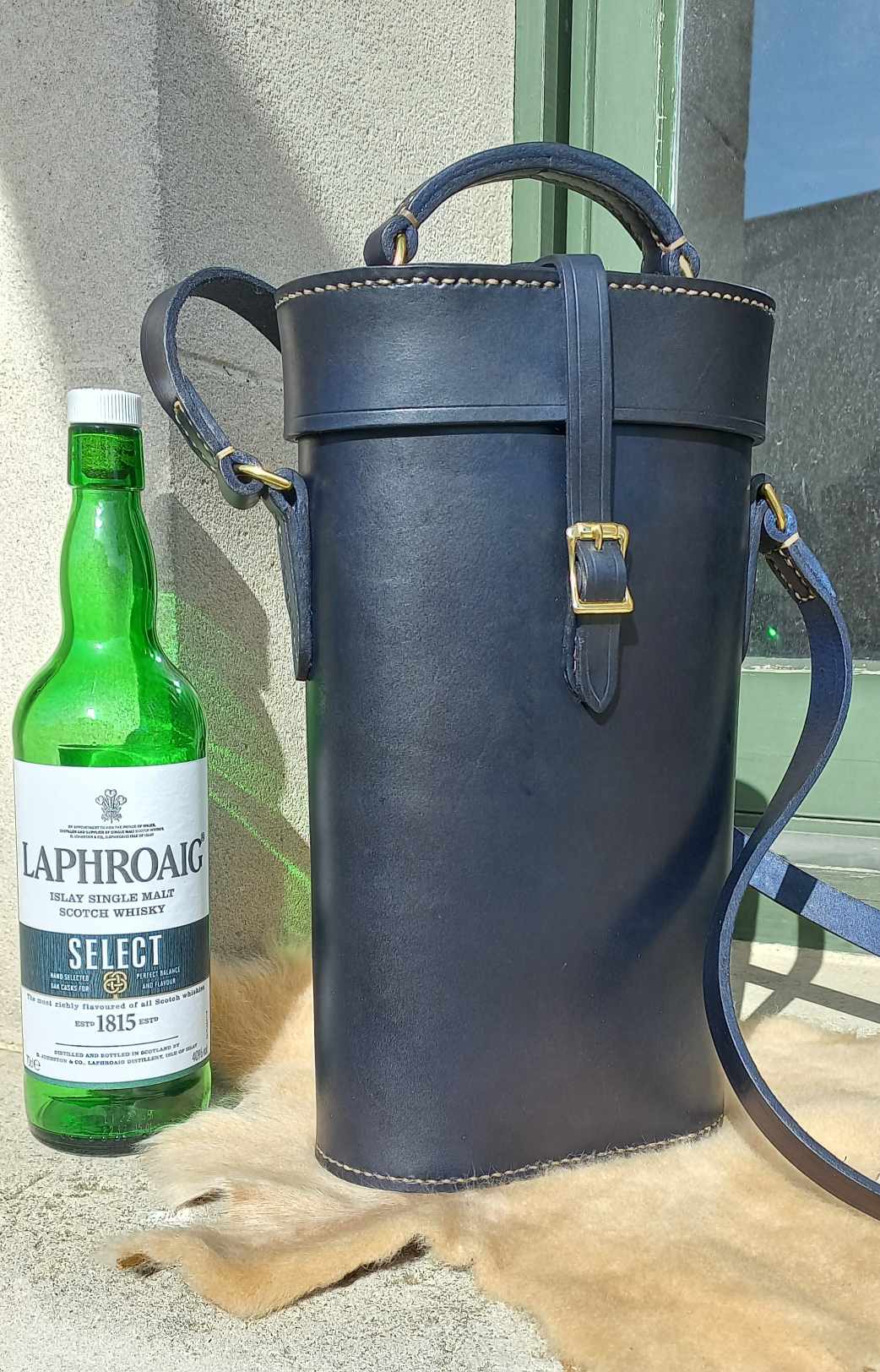 A tall case designed to carry bottles hand-made with navy blue dyed leather and gold hardware, placed on a sheepskin next to a green bottle of Scottish whisky.