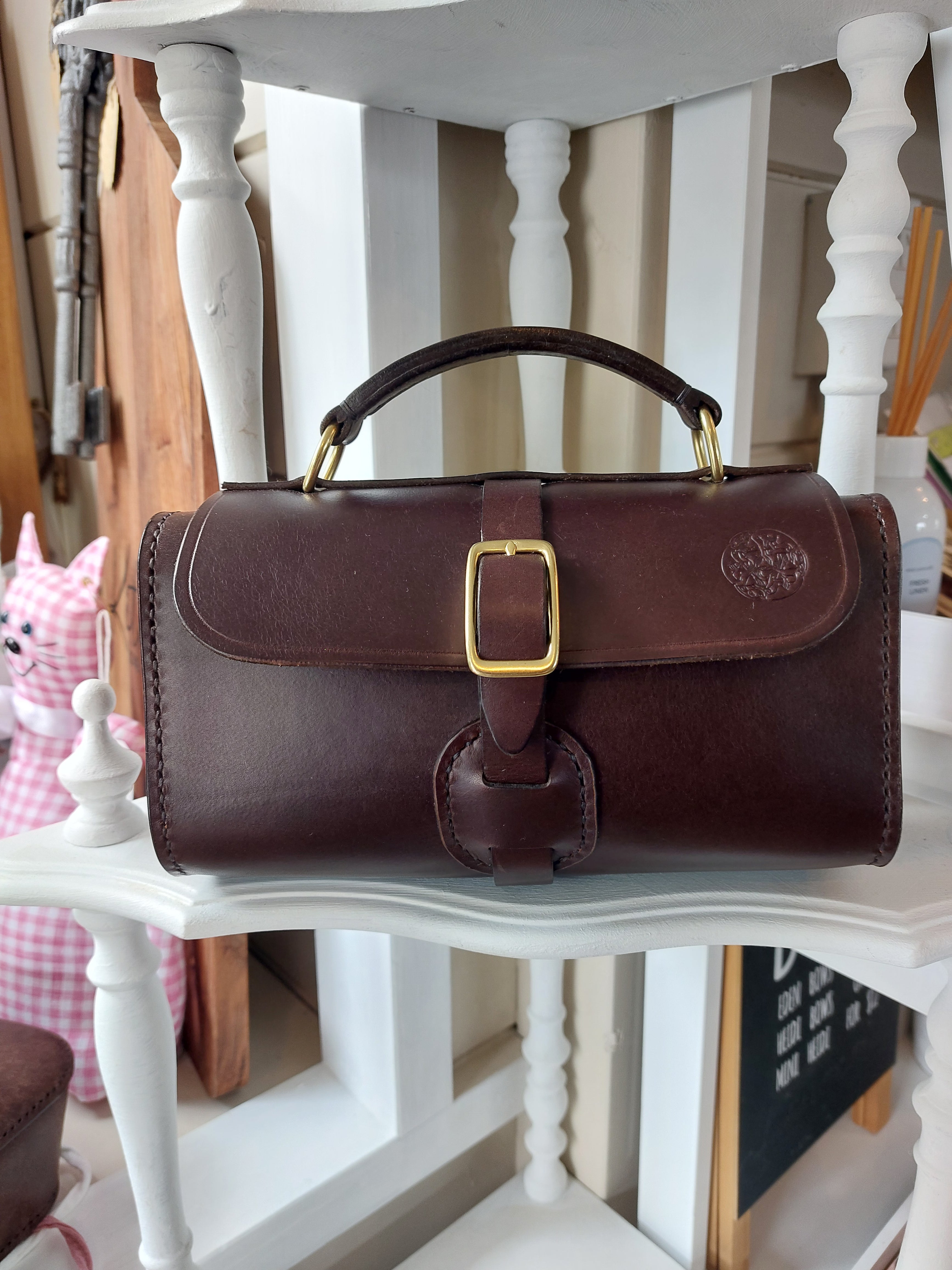 Excelsior Hand Bag made from quality Italian rich brown leather, complete with solid brass hardware, sits atop a white shelf. Stamped with the Albannach Crafts logo.