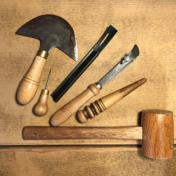 A collection of leather-working tools with wooden handles, arranged atop a wooden workshop bench.