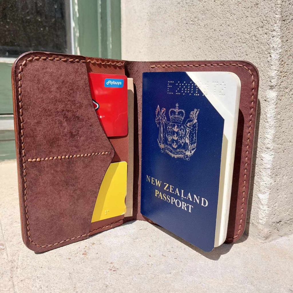 Passport Wallet made with quality brown vegetable-tanned leather, open displaying a passport and bank cards outside a historic cottage.