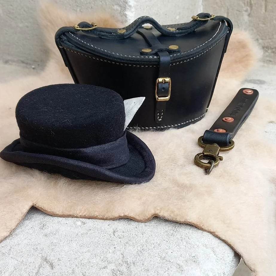 A black leather case with gold hardware next to a matching strap and old black bowler hat, placed atop a sheepskin.